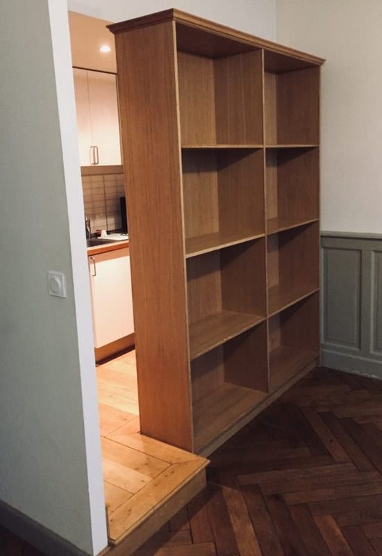 Grande bibliothèque en bois clair séparant un salon à parquet foncé d'une cuisine moderne avec sol en bois clair.