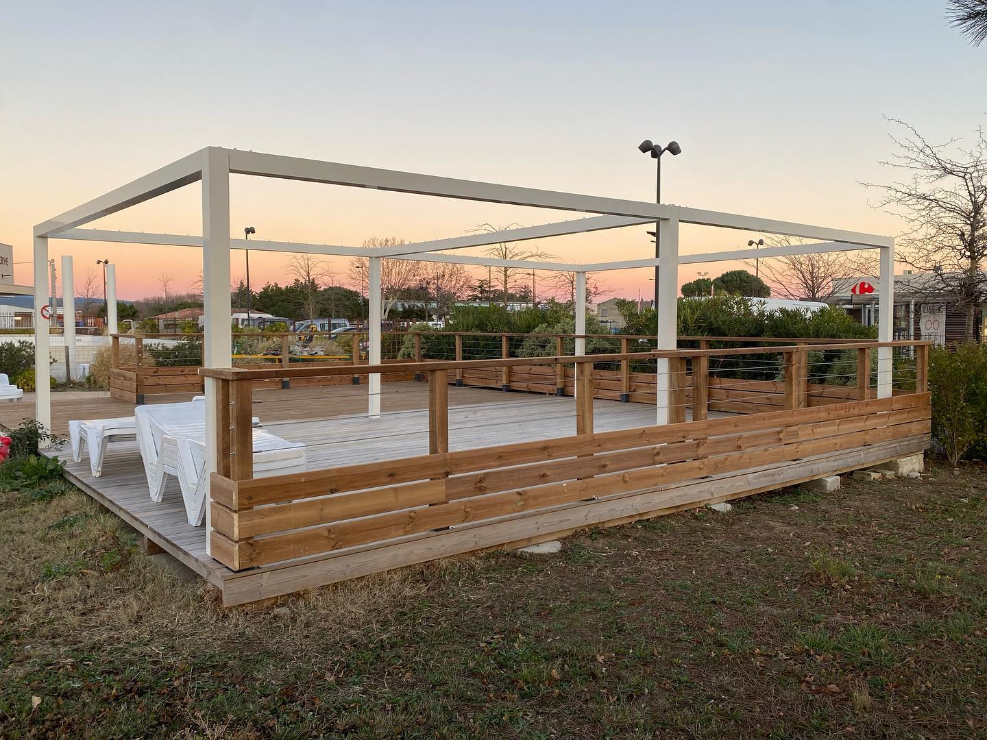 Terrasse en bois avec pergola blanche et garde-corps à câbles, au coucher du soleil, près de transats blancs.