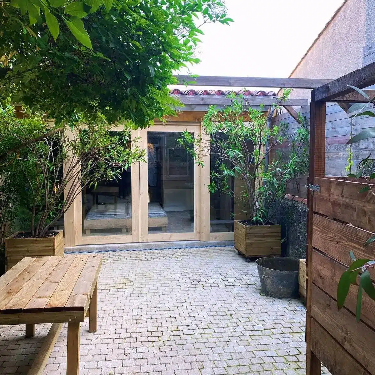 Patio extérieur avec sol pavé, table en bois, et grandes portes vitrées menant à une pièce lumineuse, entouré de verdure en bacs.
