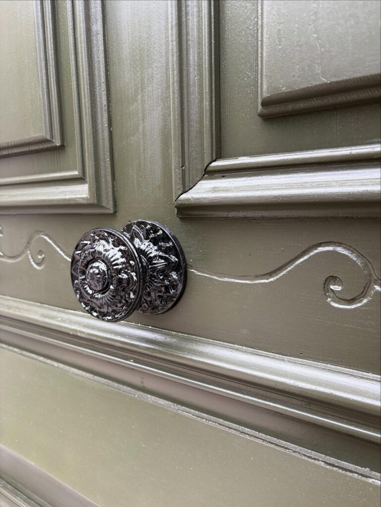 Gros plan sur une poignée de porte ancienne noire et ouvragée, fixée sur une porte en bois vert olive à panneaux moulurés.