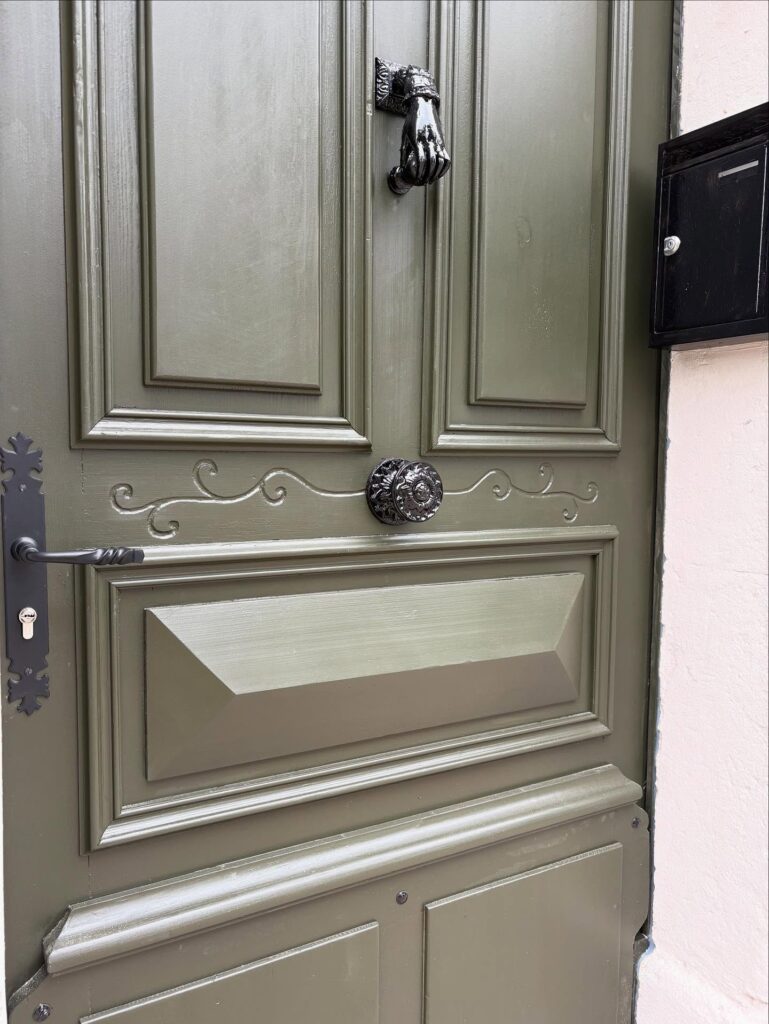 Porte en bois vert olive avec moulures, poignée noire ornementée, un heurtoir main noire, et une boîte aux lettres noire.