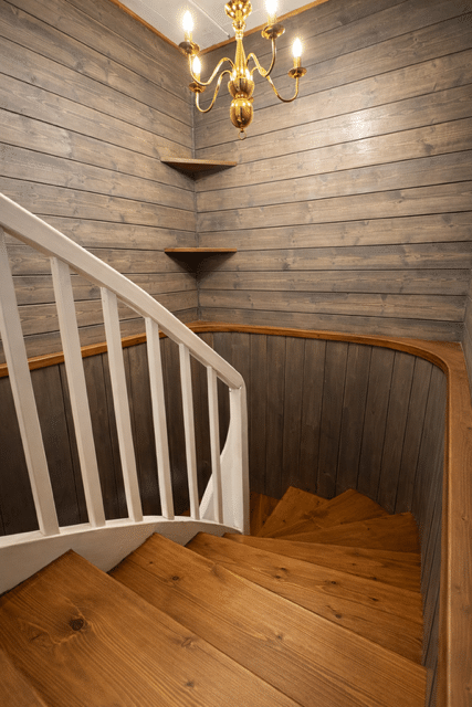 Escalier tournant en bois clair, rambarde blanche, murs en lambris gris et lustre doré suspendu.