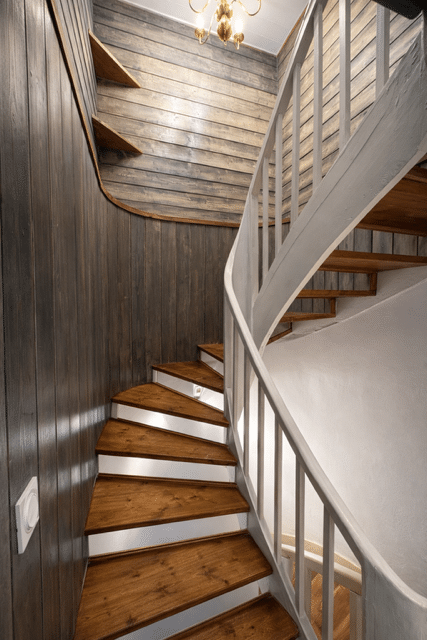 Escalier colimaçon en bois, marches chêne, risers blancs, murs lambris gris et lustre laiton doré.