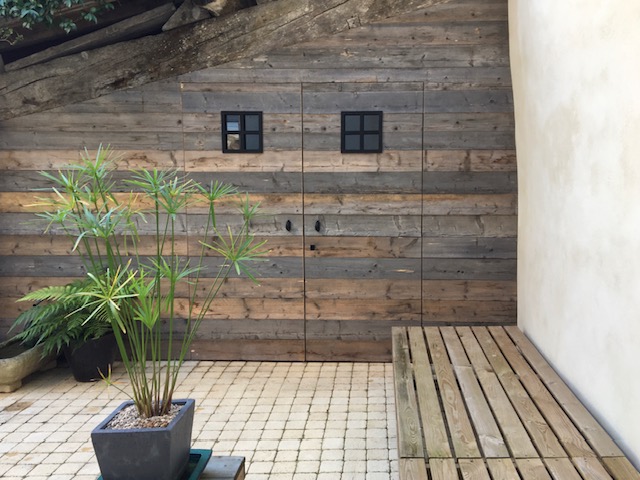 Double porte en bois vieilli, fenêtres à carreaux, plante papyrus et terrasse pavée.